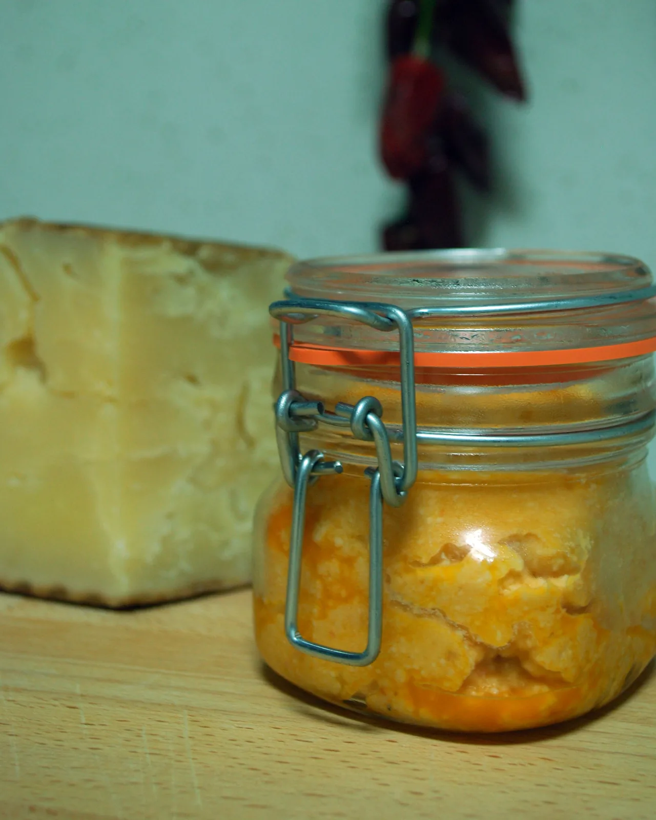 Orangefarbener, leicht körniger Almogrote in einem Bügelglas, dahinter ein Stück harter gereifter Käse und getrocknete rote Chilischoten