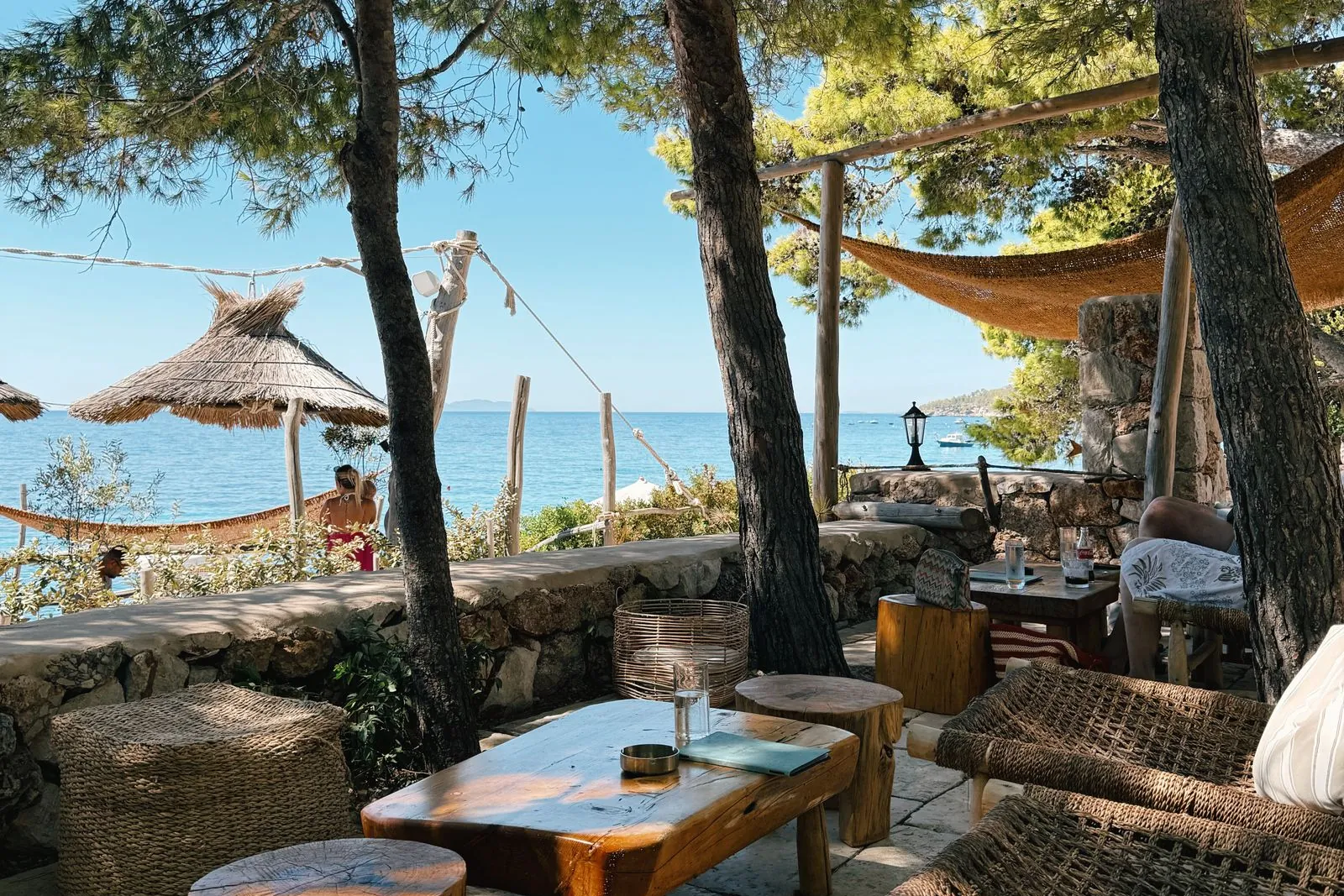 Terrasse mit niedrigen Holztischen im Schatten von Pinien, dahinter das Meer