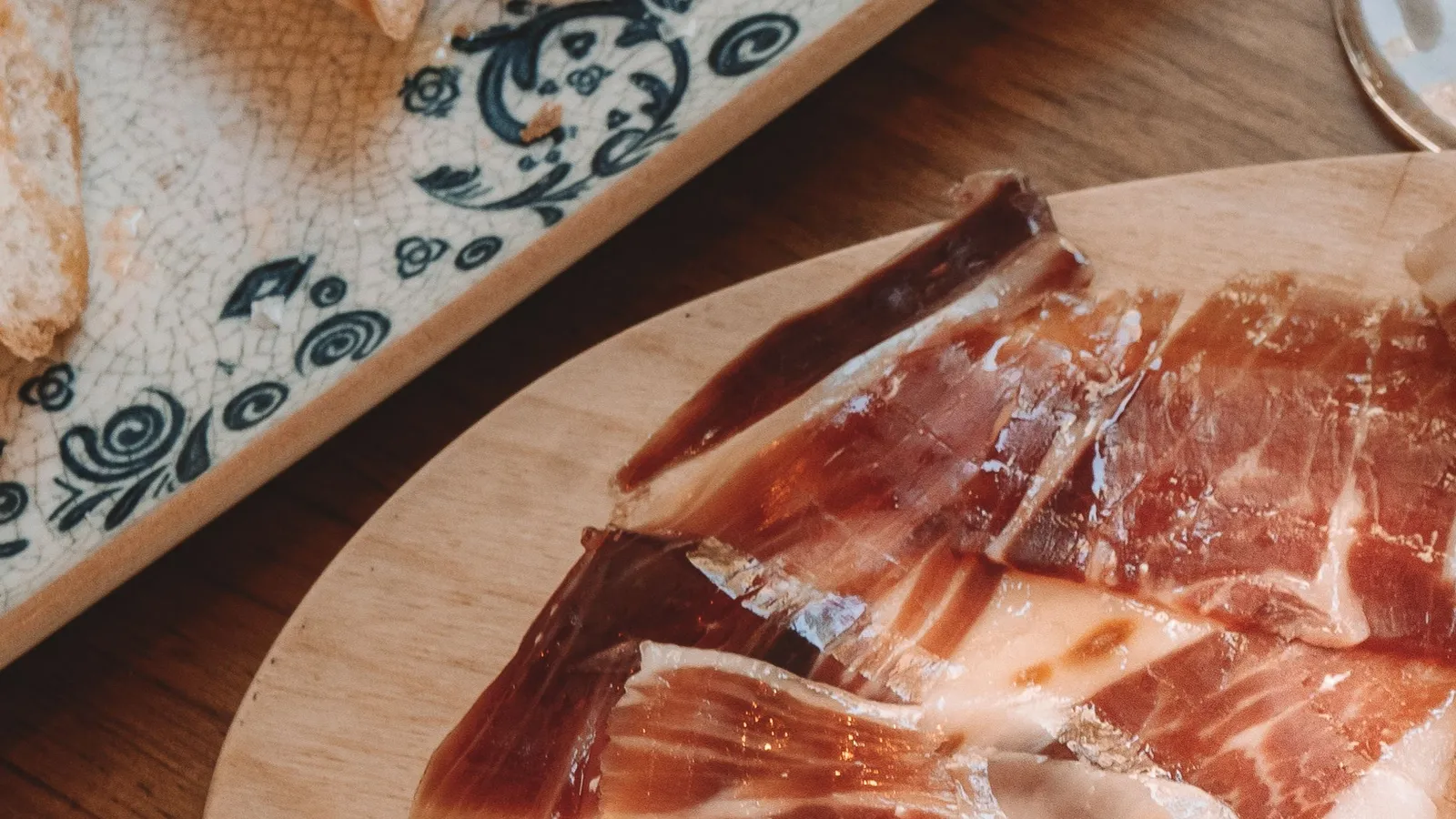 Dünn geschnittener Jamón auf einem Holzbrett, daneben Pan con tomate auf einer blau-weiß bemalten Keramikplatte