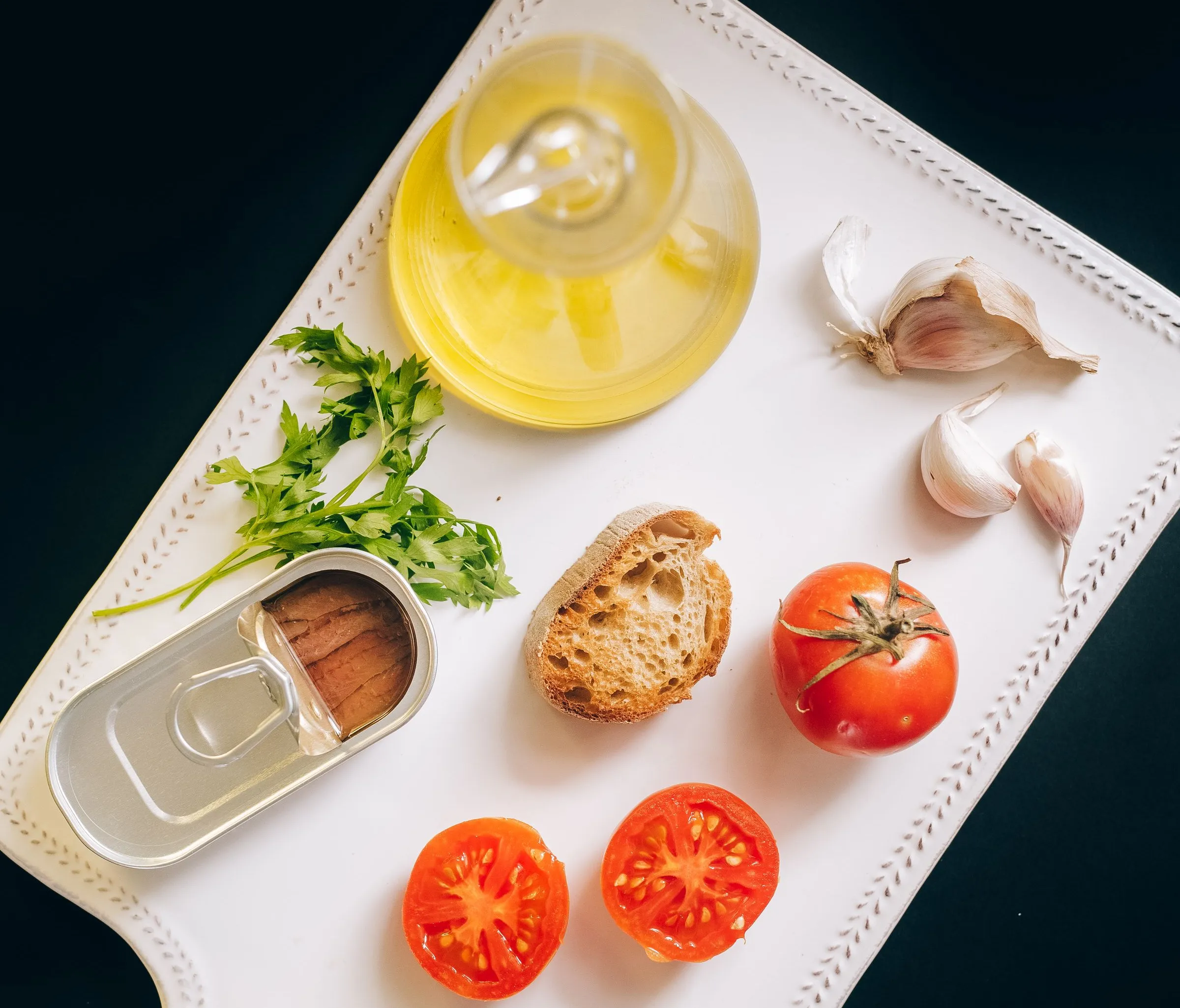Olivenöl in einer Glaskaraffe, Brot, Tomaten, Knoblauch und Sardellen auf einem weißen Brett