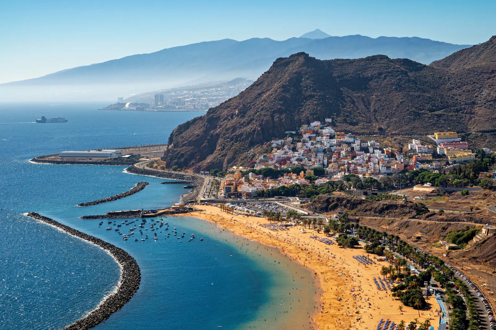 Playa de Las Teresitas bei Santa Cruz: heller Sandstrand mit Palmen, Wellenbrecher, das Fischerdorf San Andrés am Berg und in der Ferne der Teide